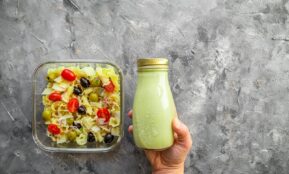 a hand holding a bottle of green smoothie next to a container of pasta salad
