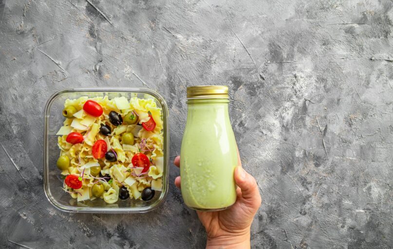 a hand holding a bottle of green smoothie next to a container of pasta salad
