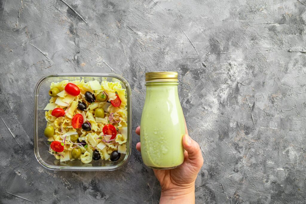 a hand holding a bottle of green smoothie next to a container of pasta salad
