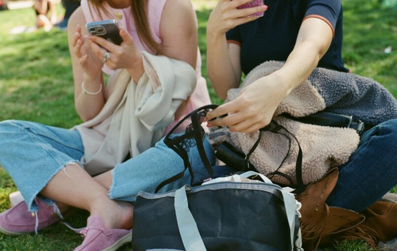 Two people sit using their phones and bags.