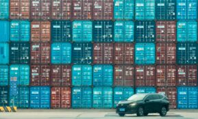 A car is parked in front of a large stack of shipping containers