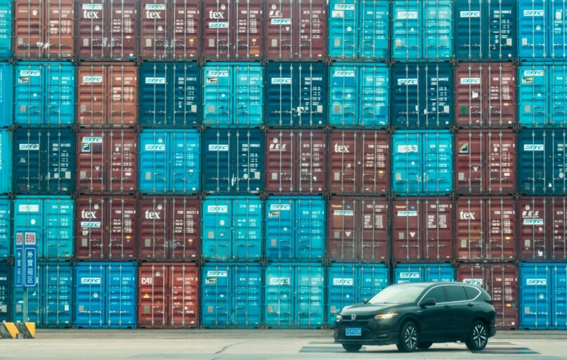 A car is parked in front of a large stack of shipping containers