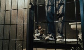 Inmate reading a book inside a prison cell while another stands nearby.