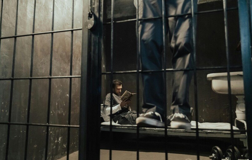 Inmate reading a book inside a prison cell while another stands nearby.