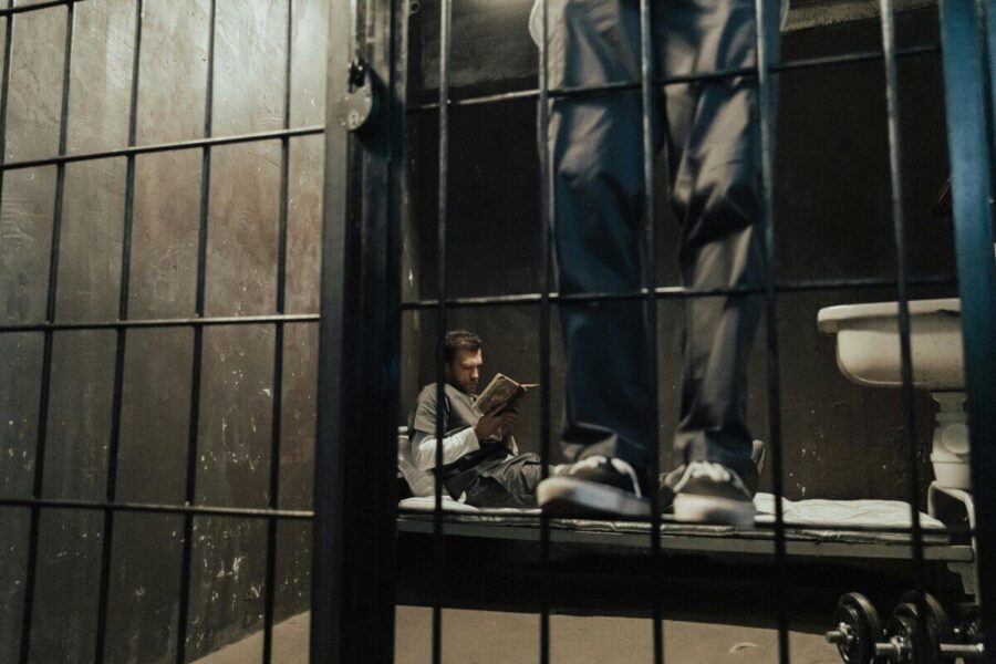 Inmate reading a book inside a prison cell while another stands nearby.