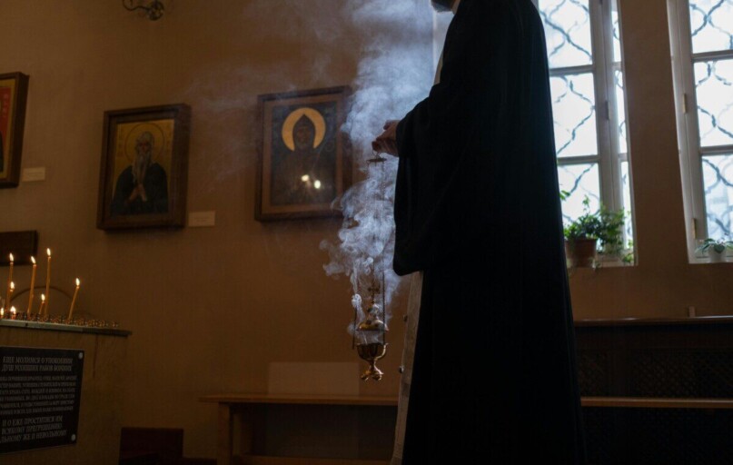 Priest holding censer with incense in a dimly lit church, surrounded by religious icons and candles.