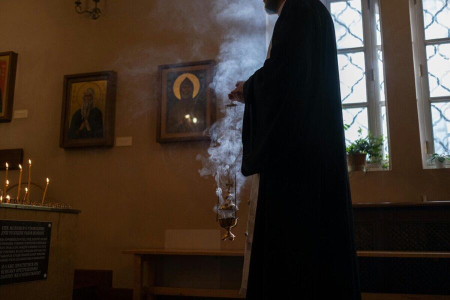 Priest holding censer with incense in a dimly lit church, surrounded by religious icons and candles.