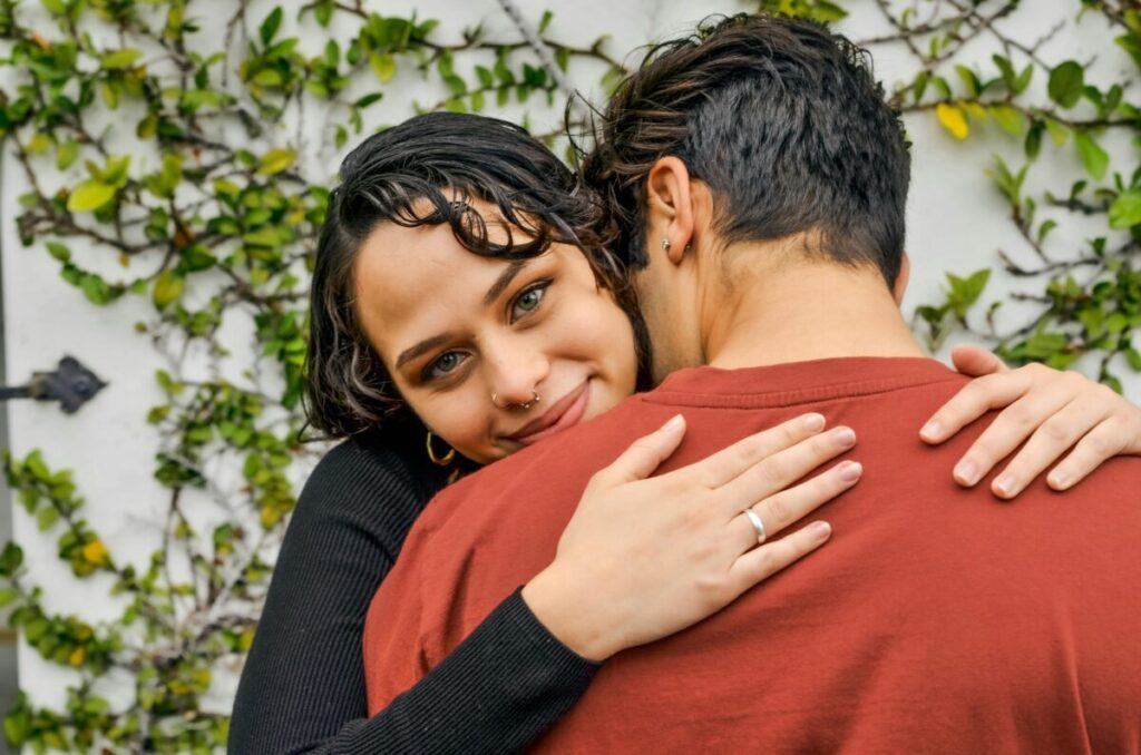 A loving couple shares a warm embrace against a leafy wall backdrop.