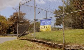 A protest banner with a "stop war" message hangs on a metal fence outdoors.
