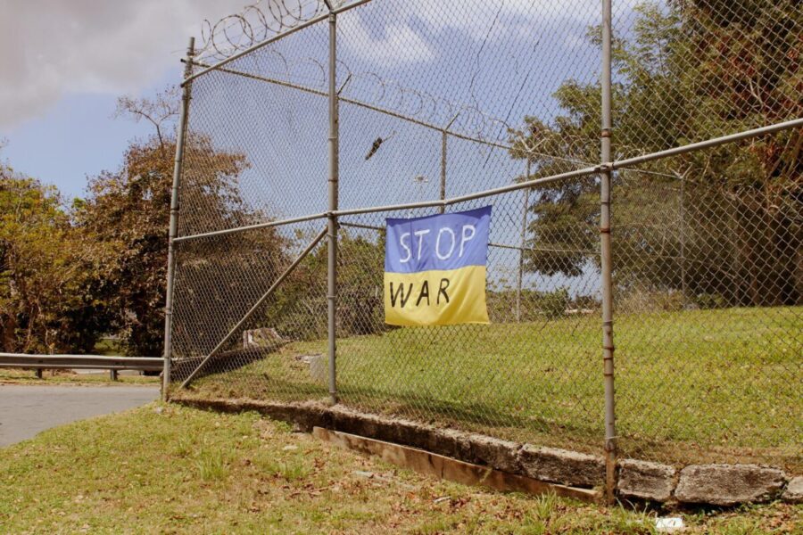 A protest banner with a "stop war" message hangs on a metal fence outdoors.