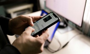 A person holding a smartphone and chatting indoors near a computer screen.