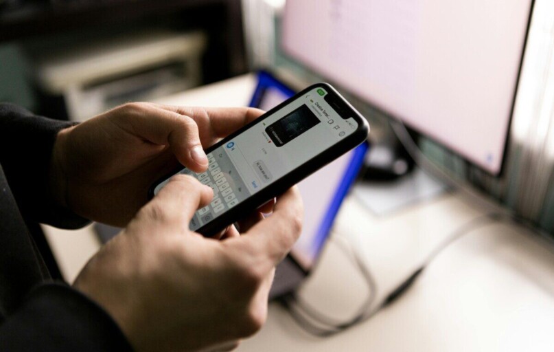 A person holding a smartphone and chatting indoors near a computer screen.