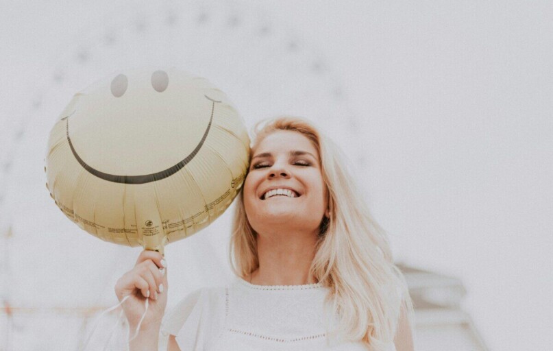 Cheerful woman holding a smiley balloon outdoors on a sunny day, exuding happiness and positivity.