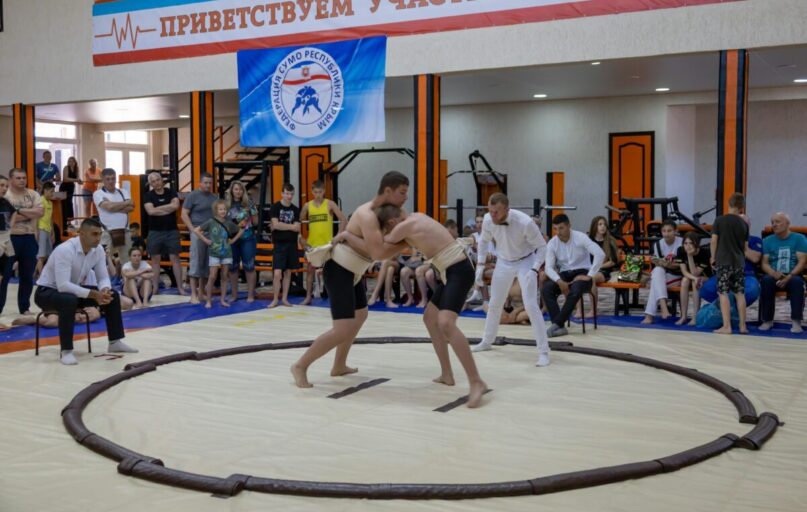 Young athletes engaged in sumo wrestling in a gym with spectators and a referee observing.