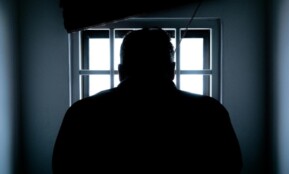 Dark silhouette of a man in a prison cell, backlit by window bars.
