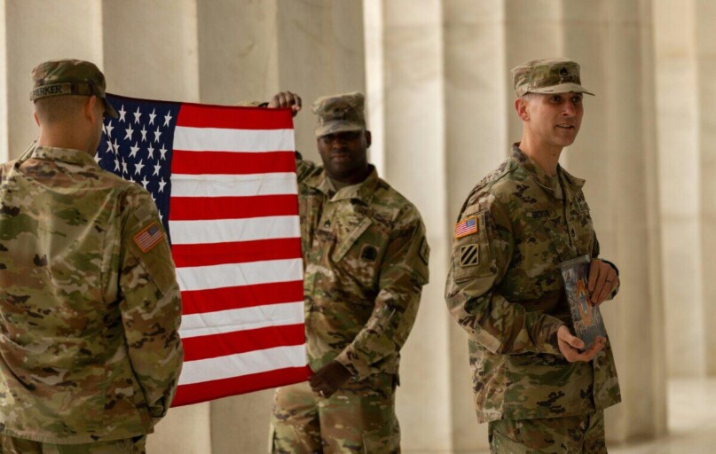 US soldiers in camouflage uniforms hold American flag at Washington DC landmark.