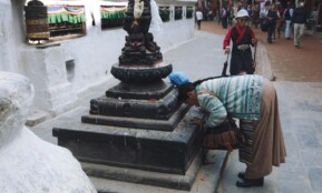 A woman in traditional attire praying at a Tibetan Buddhist stupa in a bustling street.