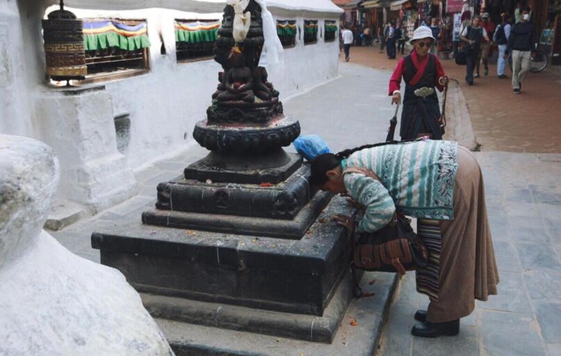 A woman in traditional attire praying at a Tibetan Buddhist stupa in a bustling street.