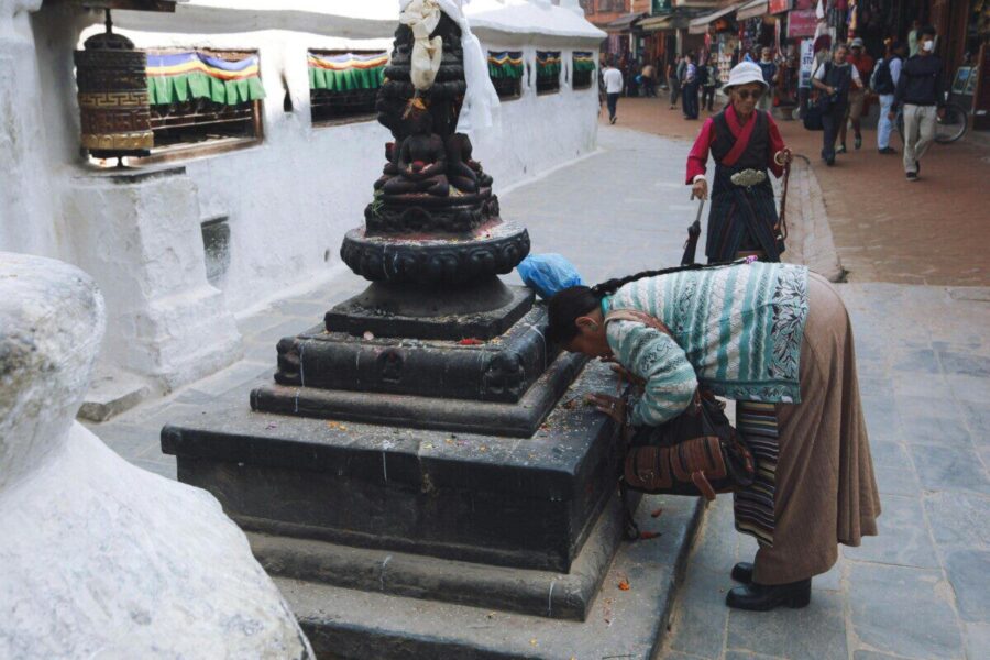 A woman in traditional attire praying at a Tibetan Buddhist stupa in a bustling street.
