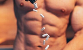 Shirtless man pouring white supplement capsules into his hand with muscles in focus.