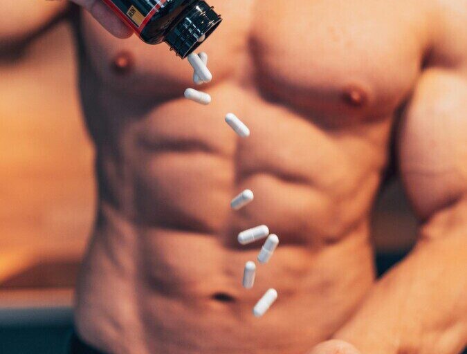 Shirtless man pouring white supplement capsules into his hand with muscles in focus.