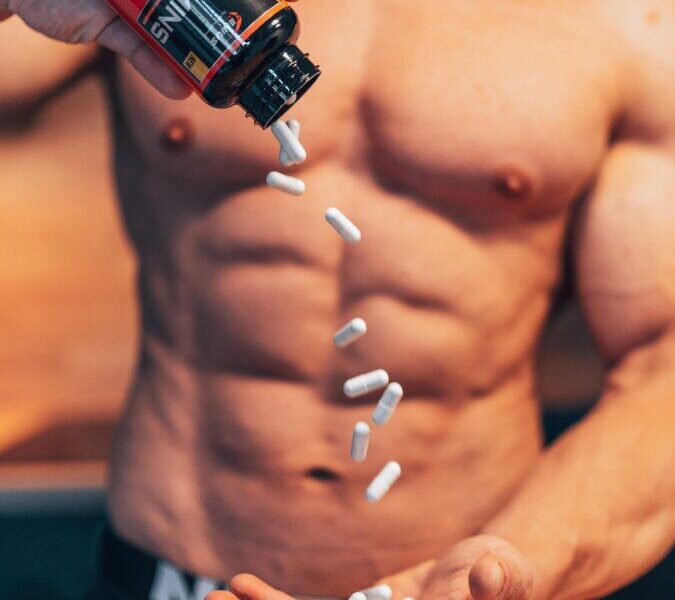 Shirtless man pouring white supplement capsules into his hand with muscles in focus.
