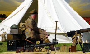 A historical reenactor dressed as a WWI soldier sits by a tent with military gear in a field.