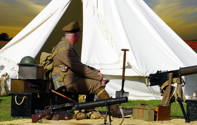 A historical reenactor dressed as a WWI soldier sits by a tent with military gear in a field.