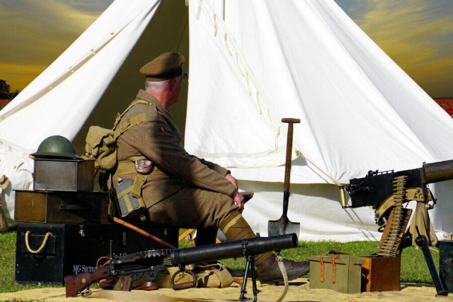 A historical reenactor dressed as a WWI soldier sits by a tent with military gear in a field.