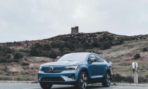 A blue Volvo electric car charging in a scenic outdoor location with a hill in the background.