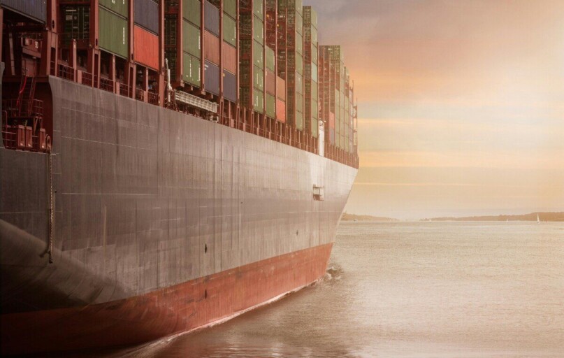 A colossal cargo ship loaded with containers navigates through calm waters against a vibrant sunset sky.