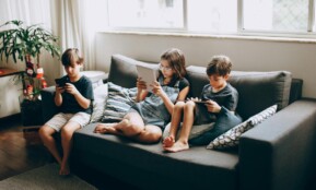 Three children on a gray sofa engrossed in digital devices, indoors.