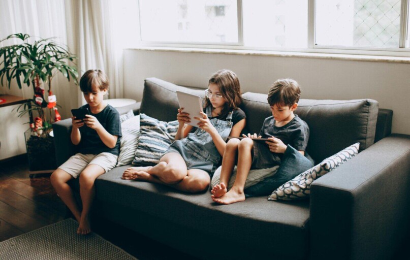 Three children on a gray sofa engrossed in digital devices, indoors.
