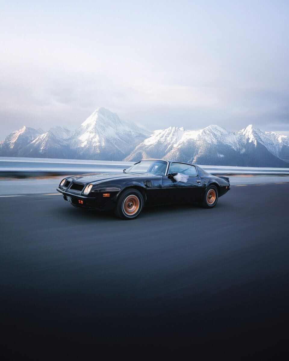 A vintage sports car speeds along a road with stunning snowy mountains in the background, exuding adventure