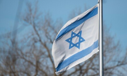 The Israeli flag featuring the Star of David against a clear blue sky and trees.