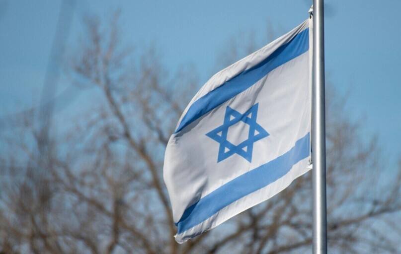 The Israeli flag featuring the Star of David against a clear blue sky and trees.