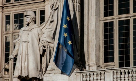 European Union flag waving beside a historic statue on stone building facade.
