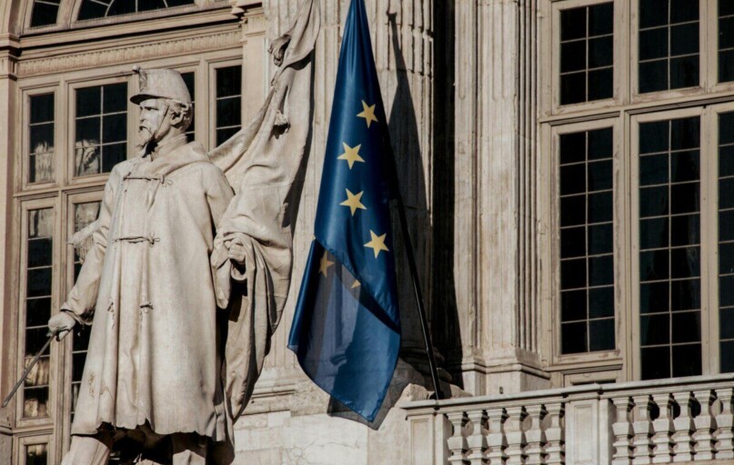 European Union flag waving beside a historic statue on stone building facade.