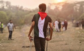 A young man wrapped in a Palestinian flag walks with crutches through a field in Bangladesh during the day.