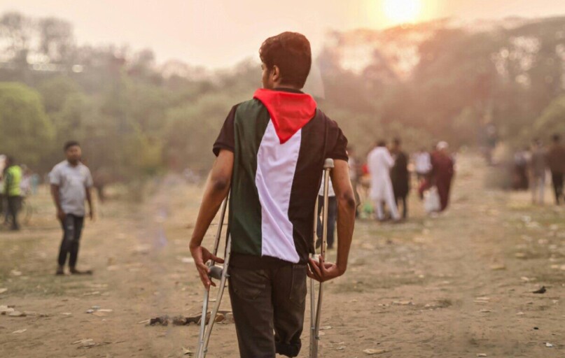 A young man wrapped in a Palestinian flag walks with crutches through a field in Bangladesh during the day.