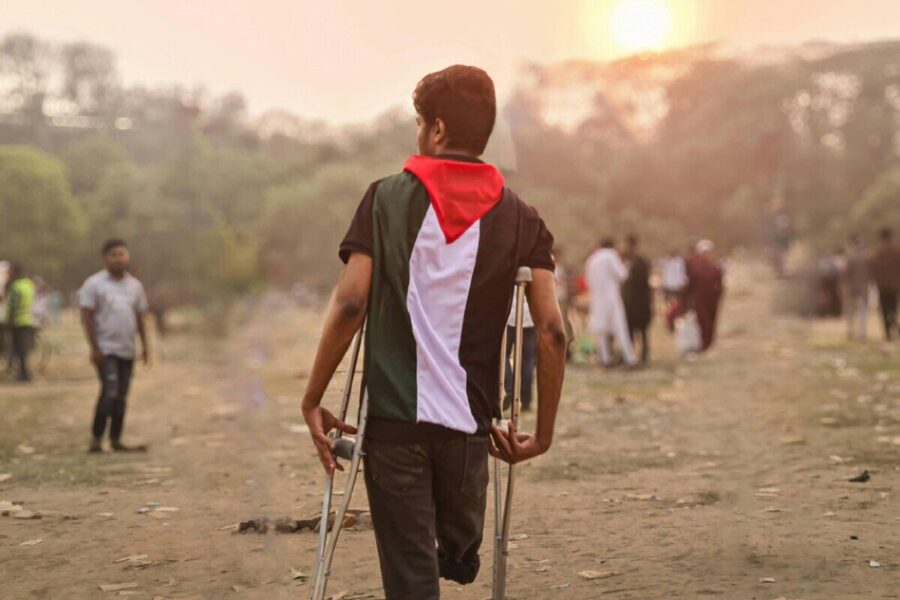 A young man wrapped in a Palestinian flag walks with crutches through a field in Bangladesh during the day.