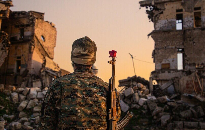 A soldier in camouflage with a red rose on rifle amidst ruined buildings at sunset.