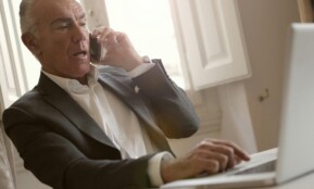 Elderly man in suit multitasking with phone and laptop in a sunlit office setting.