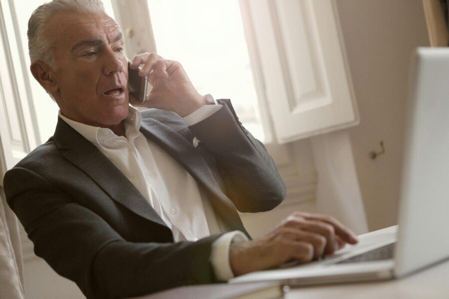 Elderly man in suit multitasking with phone and laptop in a sunlit office setting.