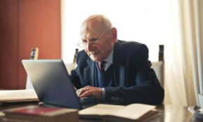 Concentrate elderly senior man in formal suit and eyeglasses working on laptop while sitting at wooden table with books in light room