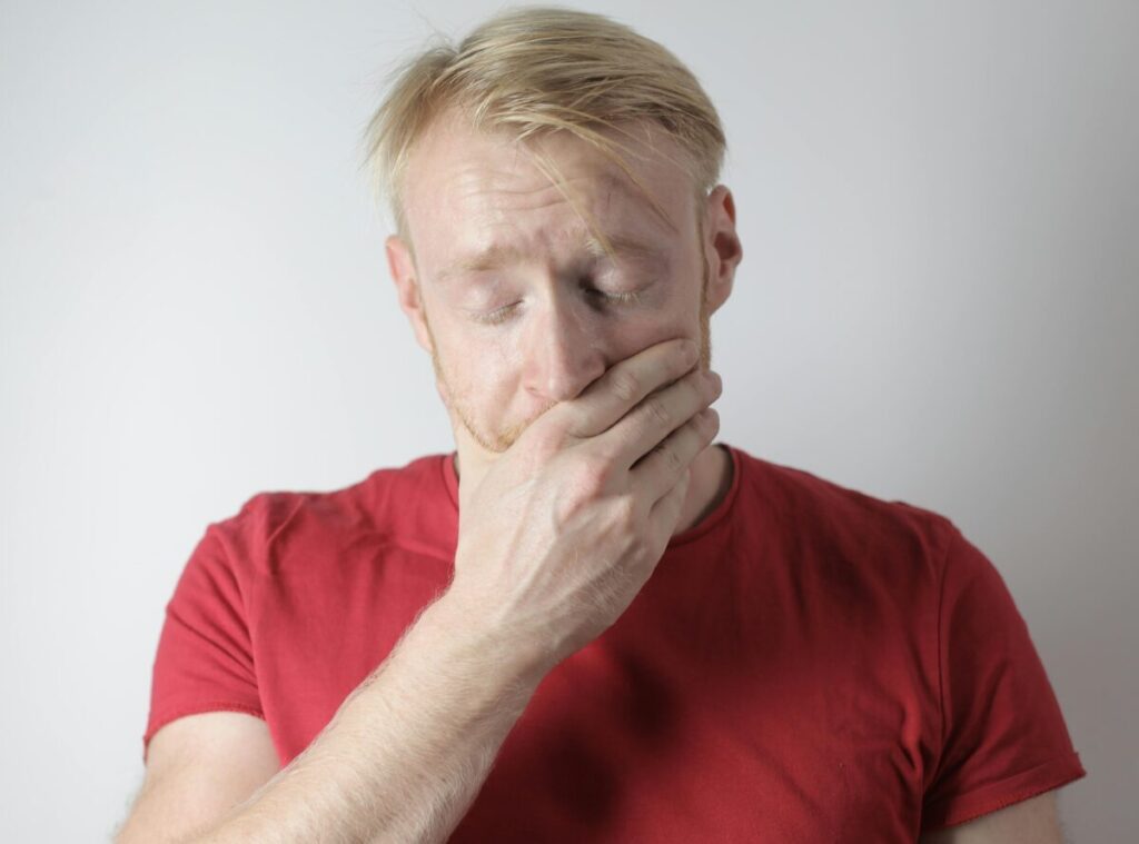 Mature man in red t shirt being in shock after getting news standing with closed eyes near gray wall in room