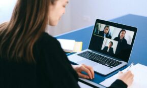 A woman engaging in a video conference using a laptop at home, taking notes.