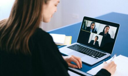 A woman engaging in a video conference using a laptop at home, taking notes.