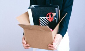 Woman carrying cardboard box with office items like scissors and notebooks.