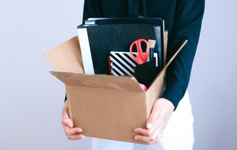 Woman carrying cardboard box with office items like scissors and notebooks.